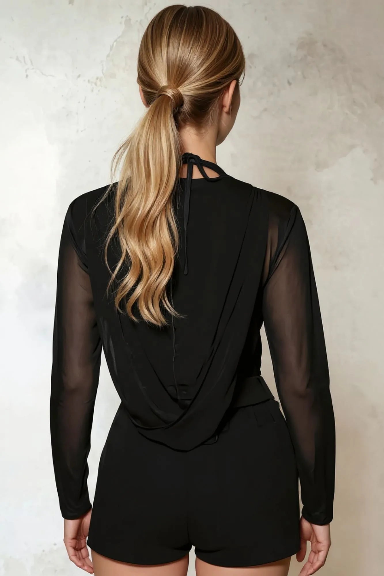 Modelo feminina vestindo blusa preta transparente em tule, com capuz, mangas longas e decote em V. Sofisticada e poderosa. Nossa blusa de tule com capuz combina transparência, decote marcante e mangas longas, criando looks irresistíveis para noites especiais.
